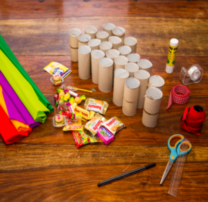 24 Leere Toilettenpapier-Rollen, 24 Süßigkeiten, Kleber, Tesafilm, eine Schnur ein Band, eine Schere, ein Lineal, ein Stift und buntes Krepp-Papier liegen auf einem Tisch.