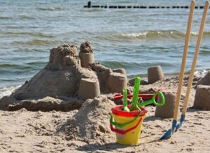 Eine Sandburg steht auf einem Strand nahe am Wasser. Daneben liegen Schippen und Eimer.