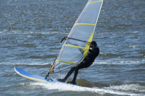 Ein Mann steht auf einem Windsurfing-Brett und hält das Segel in der Hand. Er hat einen schwarzen Neopren Anzug an.