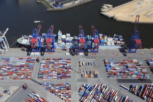 Am Hamburger Hafen entladen große Kräne Container von einem Schiff. Zahlreiche Container stehen auf dem Kai.