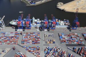 Am Hamburger Hafen entladen große Kräne Container von einem Schiff. Zahlreiche Container stehen auf dem Kai.