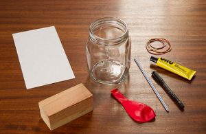 Eine Glasdose, ein Holzstück, Ein Luftballon, Kleber, ein Gummiband, ein Papier und zwei Stifte liegen auf einem Tisch.