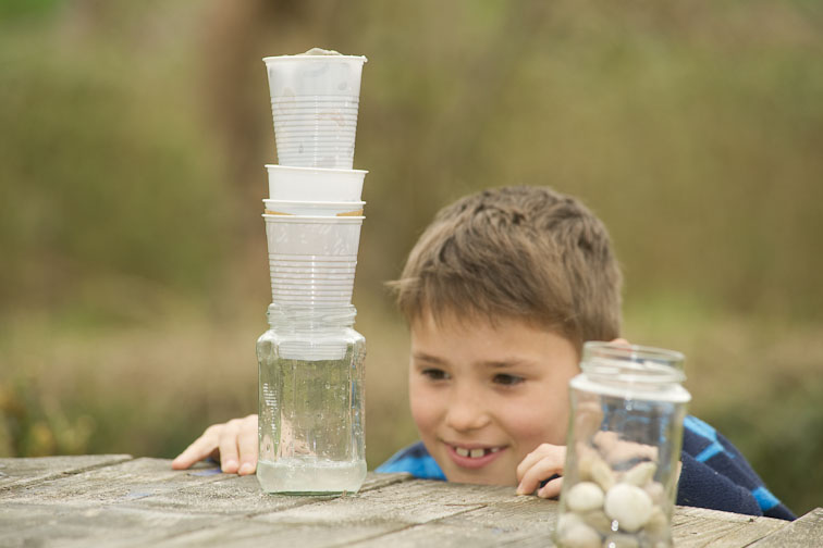 Ein Junge schaut auf einen Turm von vier übereinandergestapelten Plastikbecher. Ganz unten ist ein Glas.