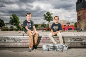 Die Jungen Louis und Bennet sitzen neben einander. Bennet sitzt auf einem Skateboard, Louis auf Stufen.