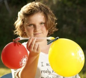 Ein Mädchen hält einen Strohhalm. Am seinem rechten und linken Ende hängt jeweils ein Luftballon.