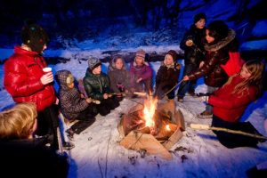 Eine Gruppe von Kindern sitzt um ein Lagerfeuer. Einige halten Stockbrot in das Feuer.
