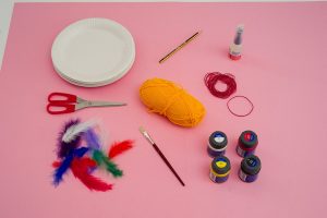 Pappteller, vier Farbtöpfe, bunte Federn, Wolle, Gummis, Schere, Kleber, ein Pinsel und ein Stift liegen auf einem Tisch.