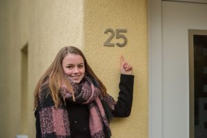 Ein Mädchen, Lotte, steht vor ihrem Haus. Sie zeigt auf die angebrachte Hausnummer.