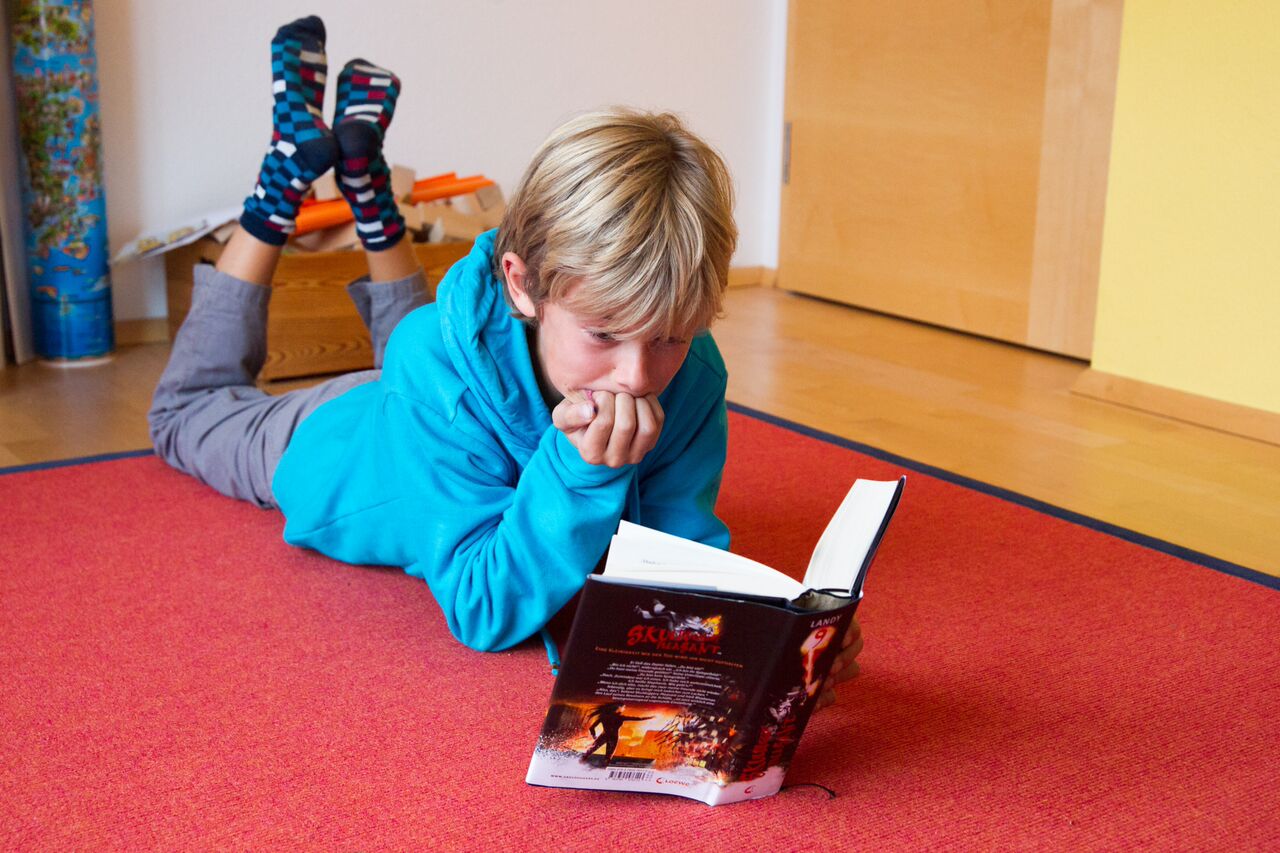 Philipp, ein blonder Junge, liegt in einem Kinderzimmer auf einem roten Teppich und liest.