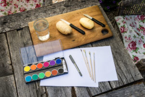 Messer, ein Brett, Pinsel, ein Glas, Papier, Stift, Farben und Kartoffeln liegen auf einem Tisch.