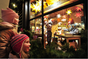 Zwei Kinder schauen durch ein Fenster auf eine Weihnachtskrippe. Dort sind zwei Zwerge in einer Küche.