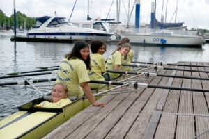 Ruderboot mit vier Kindern als Ruderer liegt am Steg an. Hinten im Boot liegt ein Mädchen. Sie ist die Steuerfrau.