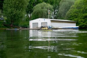 Ein Bootshaus. Vor ihm rudern vier Kinder ein Boot.