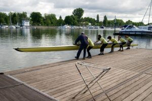 Ein Ruderer- Team und ihr Trainer legen ein Ruderboot vom Steg in das Wasser.