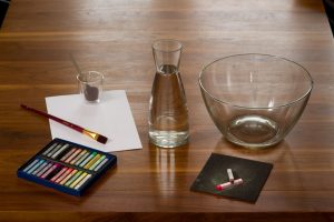 Eine Karaffe Wasser, zwei Löffel Kaffee, eine Schale ein brauner Stift, ein roter Stift, ein Papier und ein Pinsel liegen auf einem Tisch.