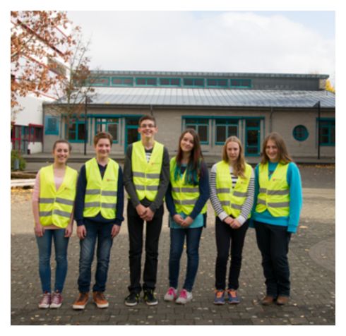 Sechs Kinder stehen auf einem Pausenhof in gelben Westen nebeneinander. Das sind die Schul-Sanitäter.