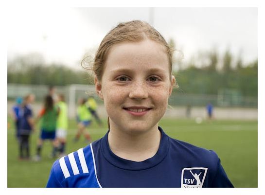 Ein Mädchen im Fußballtrikot schaut uns an. Sie steht auf einem Fußballfeld.  