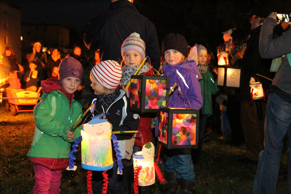 Ein Sankt-Martins-Umzug mit Laternen bei Nacht. Vor uns stehen vier Kinder mit bunt geschmückten Laternen.
