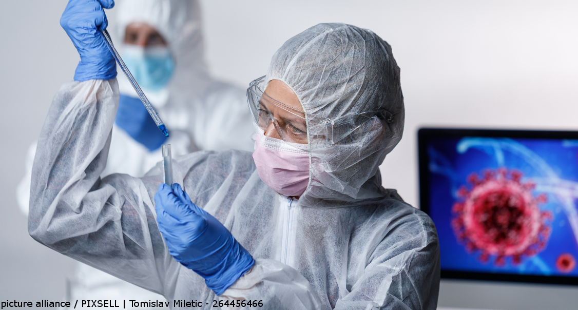 Eine Ärztin in Kroatien in Schutzkleidung, mit Mundschutz und Schutzbrille, träufelt einige Tropfen in ein Reagenzglas. Hinter ihr steht ein Bildschirm, auf dem ein roter Virus zu sehen ist.