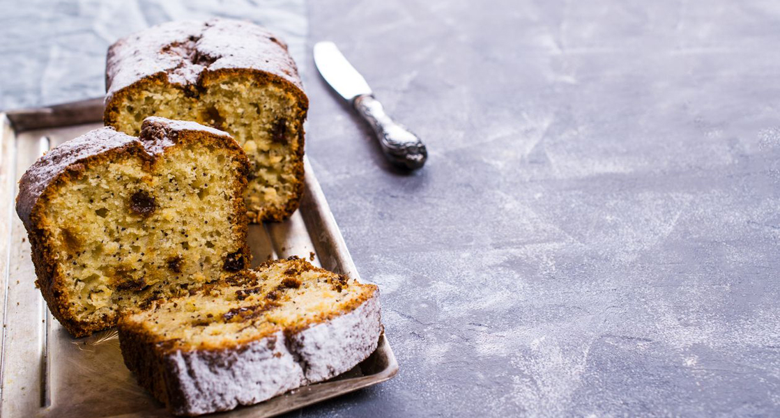 Ein Kastenkuchen mit Rosinen steht auf einem Tisch. Von ihm sind zwei Scheiben abgeschnitten. Ein Messer liegt rechts daneben.