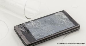 Ein schwarzes Smartphone liegt mit einem zersplitterten Bildschirm auf einem Tisch. Links daneben ist ein Glas umgekippt. Das Wasser daraus ist auf das Handy gelaufen.