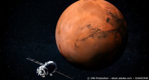 Der Mars, der rote Planet. Neben ihm fliegt ein Satellit.