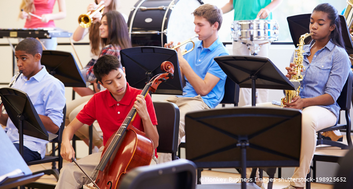 Mehrere Schulkinder spielen zusammen ihre Musikinstrumente: Ein Cello, ein Saxofon, eine Trommel, eine Geige und zwei Trompeten.
