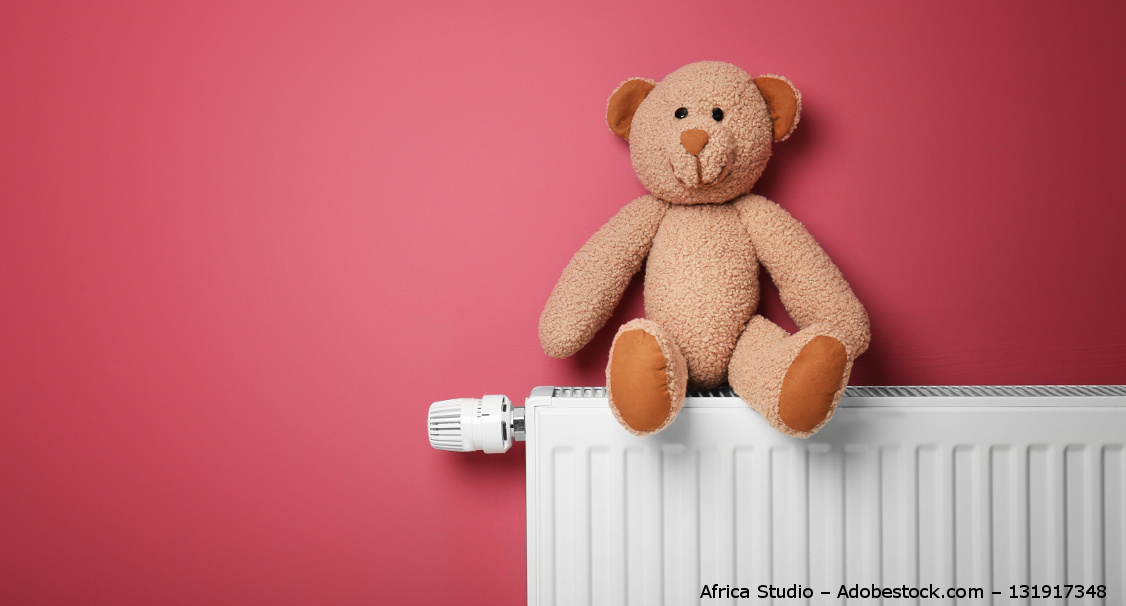 Ein brauner Teddy sitzt auf einer Heizung. Hinter ihm ist eine Wand in rosa.