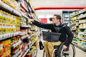 Gleiche Chancen zählen: Ein junger Mann der im Rollstuhl sitzt kauft in einem Supermarkt ein. Er steht vor einem Regal und nimmt eine Dose heraus, der Einkaufskorb steht auf seinem Schoß.