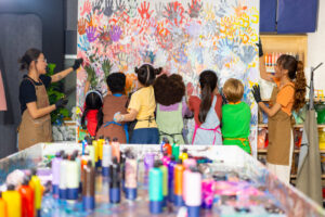 Eine Gruppe von verschiedenen Schulkindern malen mit ihren Händen ein Bild. Die Kunst findet auf Leinwand einer Leinwand Platz. Im Kunststudio sind glücklich aussehende Jungen und Mädchen, die rechts und links von zwei jungen Lehrerinnen begleitet werden. Im Vordergrund sieht man viele Farbtuben.